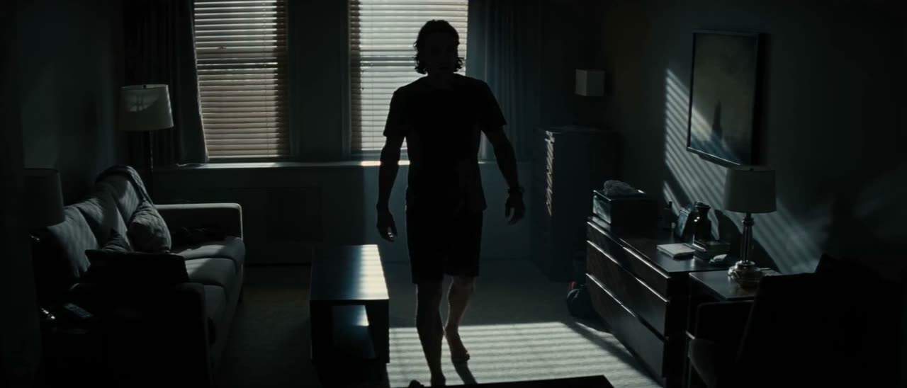 A silhouetted figure crossing a dark bedroom striped with light through blinds